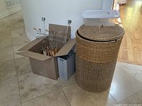 Wooden hangers inside a cardboard box, metal over-door clothes rack placed on top, grey drawer organizers stacked beside box, and two-tone woven laundry basket with lid.
