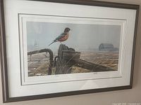 Framed print showing a robin perched on a weathered wooden fence post in a farm field with barn buildings in the distance.