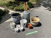 Full view of assorted garden pots of various materials and sizes arranged outdoors on pavement including a large grey metal decorative pot, a tall grey ceramic pot, a large beige pot, smaller white pots, a small terracotta pot, and a glass pot.