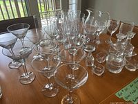 Various clear glassware set on wooden table including two sizes of martini glasses, pilsner glasses, a large pitcher, oil and vinegar set, wine glasses, candle holders and small glass bowls