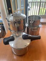 Photo of a Lagostina stainless steel pressure cooker with black detachable handle and locking lid, with a large stock pot, two medium pots and two perforated steamer inserts stacked next to it on a wooden surface.