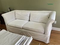White canvas upholstered sofa with two loose seat and back cushions, sitting on hardwood floor against a green wall.