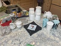 Photo showing a variety of ceramic and glass items including white ceramic teapot, white lidded mugs, cream and sugar set, colorful mugs, glass condiment dish, and watermelon napkin holder on kitchen counter.