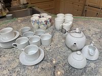 Wide view of assortment showing white porcelain cups, mugs, saucers, floral mugs, ribbed mugs, and white teapot with metal infuser, sugar bowl, and creamer.
