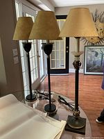 Photo showing the three lamps from a distance on a glass table with beige lampshades and visible cords.