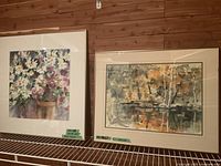 Two framed watercolour paintings side by side on a metal shelf with wooden wall background. The left is a floral scene and the right is a landscape with trees and water, both framed and matted.
