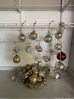 Photo showing a variety of Christmas ball ornaments hanging from hooks against a pegboard and some gathered in a pile with netted gold string at the bottom.