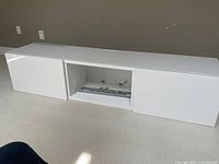 White wall mounted storage cabinet with glass top seen from front, closed doors, with central open shelf holding metal hardware.