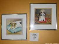 Photograph showing two framed watercolours featuring dogs, placed against a beige wall.