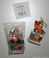 Two A&W Root Beer Christmas ornaments shown side by side; one still in original packaging, one displayed showing A&W bear in orange sweater sitting on chimney with fire.