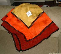 Folded hand crocheted afghan draped over a couch, showing ochre, orange, and rust color blocks with brown border.