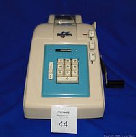 Front view of vintage J Regna manual calculator showing turquoise numbered keypad and cream plastic body with crank handle on right side.