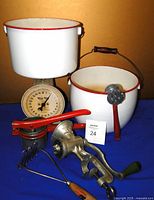 Front view showing white enamel pots with red rims, wooden handle on one pot, vintage Way-Rite country scale with cream face and black numbers, manual meat grinder, and red-handled kitchen utensil.