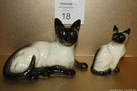 Front view of two Siamese cat figurines, larger lying down and smaller sitting upright, showing detailed paint and facial features.