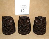 Front view of three cast iron owl votive candle holders standing side by side on a beige surface with auction lot number in background.
