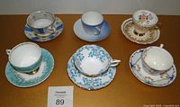 Front view of all six tea cup and saucer sets arranged in two rows on a table, showing decorative patterns and colors.