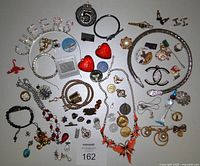 Top-down view of assorted jewelry, pins, tokens, and accessories arranged on a neutral surface including chains, bracelets, and pins.