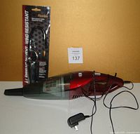 Photo showing the red and black rechargeable mini handheld vacuum with transparent dust container, power switch, and charging adapter with cord.