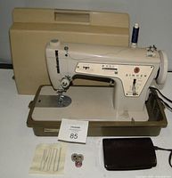 Full view of the Singer sewing machine with beige hard carrying case behind it, including bobbins and stitch sample fabric in front.
