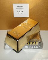 One gold bar style door stop shown outside of packaging with embossed gold bar markings on top and MaxSold auction lot number in the background.