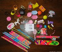 Full overview of all items laid on a wooden surface, showing skis, tennis rackets, small basketballs, Barbie sun visors, white poodle figurine, miniature cookware, and various small plastic accessories.