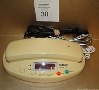 Front view of the phone base showing the cream color, digital clock display, buttons for setting time, alarm and snooze, and included cords.