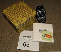 Vintage Fossil yellow geometric patterned metal storage box, watch with black square dial and leather bund band, instruction manual, and branded sticker, all laid out on carpet.