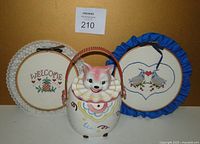 Photo of the kitten cookie jar with handle and two framed cross stitch pieces on a beige background.
