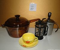 Photo shows one brown translucent Visions cookware pot with lid, two metal coffee presses, and a yellow ceramic cup and saucer with cherry decoration.