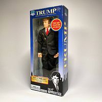 Front view of the boxed Donald J. Trump talking doll from 2004 The Apprentice series with blue packaging showing the doll wearing a dark suit and red tie.