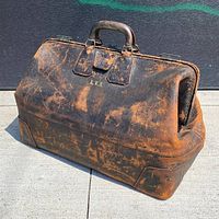 Front angled view of the large antique travel bag. Shows distressed leather with metal clasps, stitched handles, and monogram LVS on front.