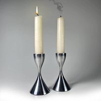 Two aluminum candleholders shown side by side, one with a lit white taper candle, the other with a smoked-out candle. Reflective brushed aluminum finish with MCM-style hourglass shape.