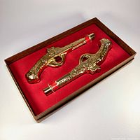 Set of two gold-tone glass dueling pistol-shaped Cologne bottles in original brown box with red lining.