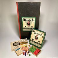 Photo showing the original Monopoly Popular Edition box with black and red exterior, foldable game board leaning against the box, colorful wooden player tokens, paper money, cards including Chance and Community Chest, two dice, and the wooden hotels and houses organized in a wooden tray.