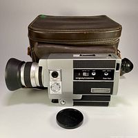 Side view of Argus/Cosina Super 8mm film movie camera with attached rubber eyepiece, lens, and black lens cap in front, brown carrying bag behind.