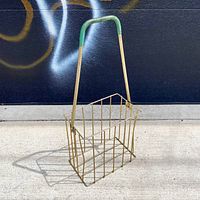 Front angle view of the vintage metal baseball carrier basket with green handle standing on concrete ground against graffiti wall background.