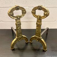 Front angle view of two brass fireplace andirons showing their oval loop tops with riveted design and arched bases with three feet each.