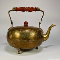 Side view of the vintage brass teapot showing the floral etching, wooden handle, three legs and gooseneck spout.