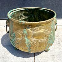 Top and side view showing the brass jardiniere planter with embossed fruit design, lion face handles, and patina on both interior and exterior.