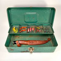 Open green metal tackle box showing 11 fishing lures and knife with leather sheath inside divided compartments