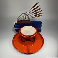 Photo showing complete fondue set: orange divided tray, ceramic bowl on black metal stand, and 6 color-coded rosewood and stainless steel forks displayed on their wooden stand with blue box behind