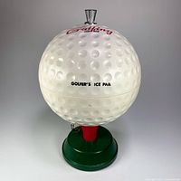 White golf ball shaped insulated ice bucket with dimpled surface and red text, silver top knob and RC logo on top, sitting on green base with red holder