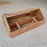 View of vintage wooden tool caddy showing four compartments and central handle, resting on concrete floor.
