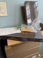 Three dark paulownia wood floating shelves on a cardboard box. Front shelf is rectangular. A small plastic bag with mounting screws and wall plugs is visible along with an instruction sheet.