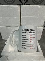Photo showing four blocks of foam packaging and one white ceramic mug in the front. Mug bears printed text 'Retirement Weekly Schedule' with days of week and humorous activities.
