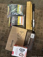 Photo showing boxed starter motor, Compustar remote starter box, Toyota dash cam hardware packaged and wrapped, and a metal engine puller tool with handle and grip.