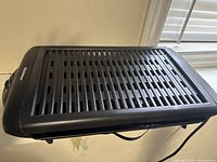 Top view of the black electric grill showing the slotted grilling surface and part of the electrical cord.
