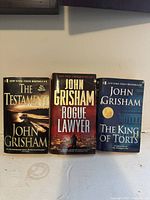Front covers of three John Grisham paperback novels: The Testament, Rogue Lawyer, The King of Torts displayed upright on a flat surface.