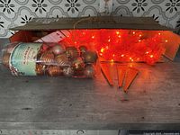 Orange illuminated artificial Christmas tree with three orange plastic stand legs and a pack of assorted orange and bronze Christmas ornaments on a table.