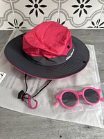 Bright pink and grey toddler sun hat with wide brim and adjustable chin strap, next to packaged pink sunglasses